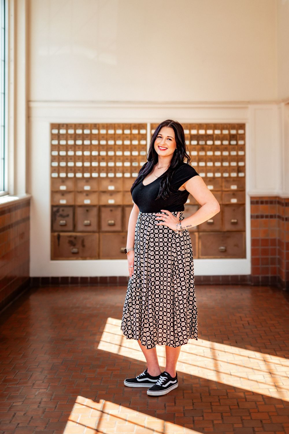 Woman in black top and patterned skirt poses in front of vintage card catalog drawers as sunlight streams through window.