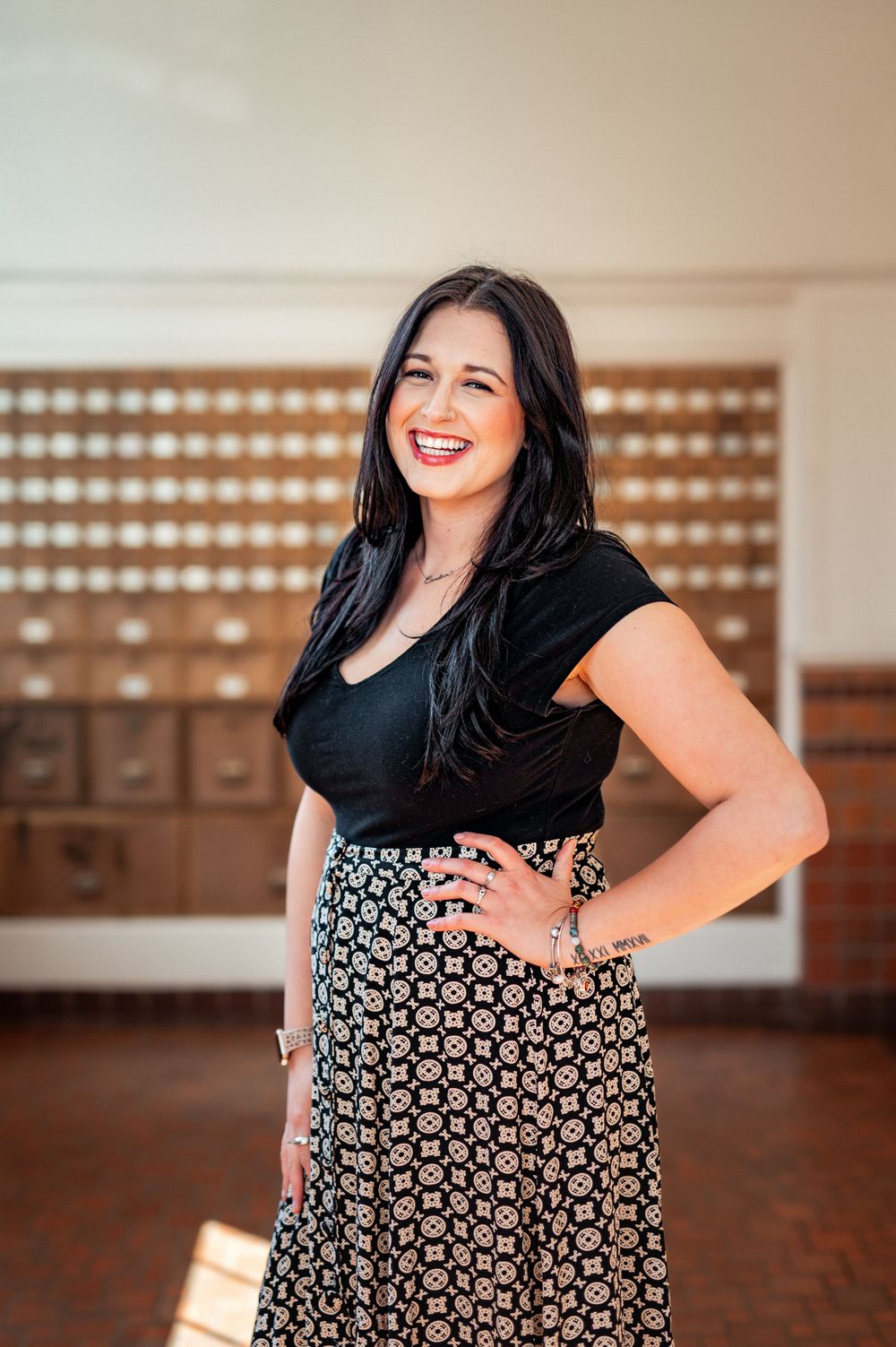 A joyful woman in black top and patterned skirt poses with hands on hips in a warm-lit indoor setting.