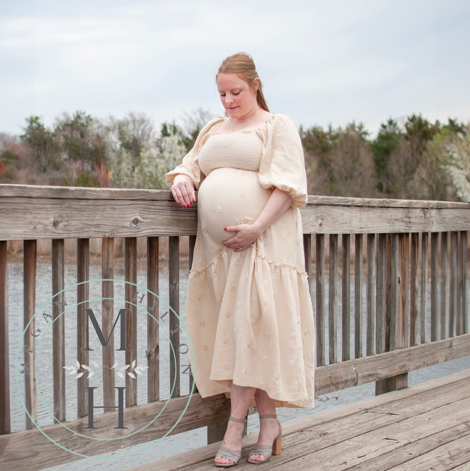 Expecting woman in cream-colored dress stands thoughtfully on wooden deck during outdoor maternity photoshoot.
