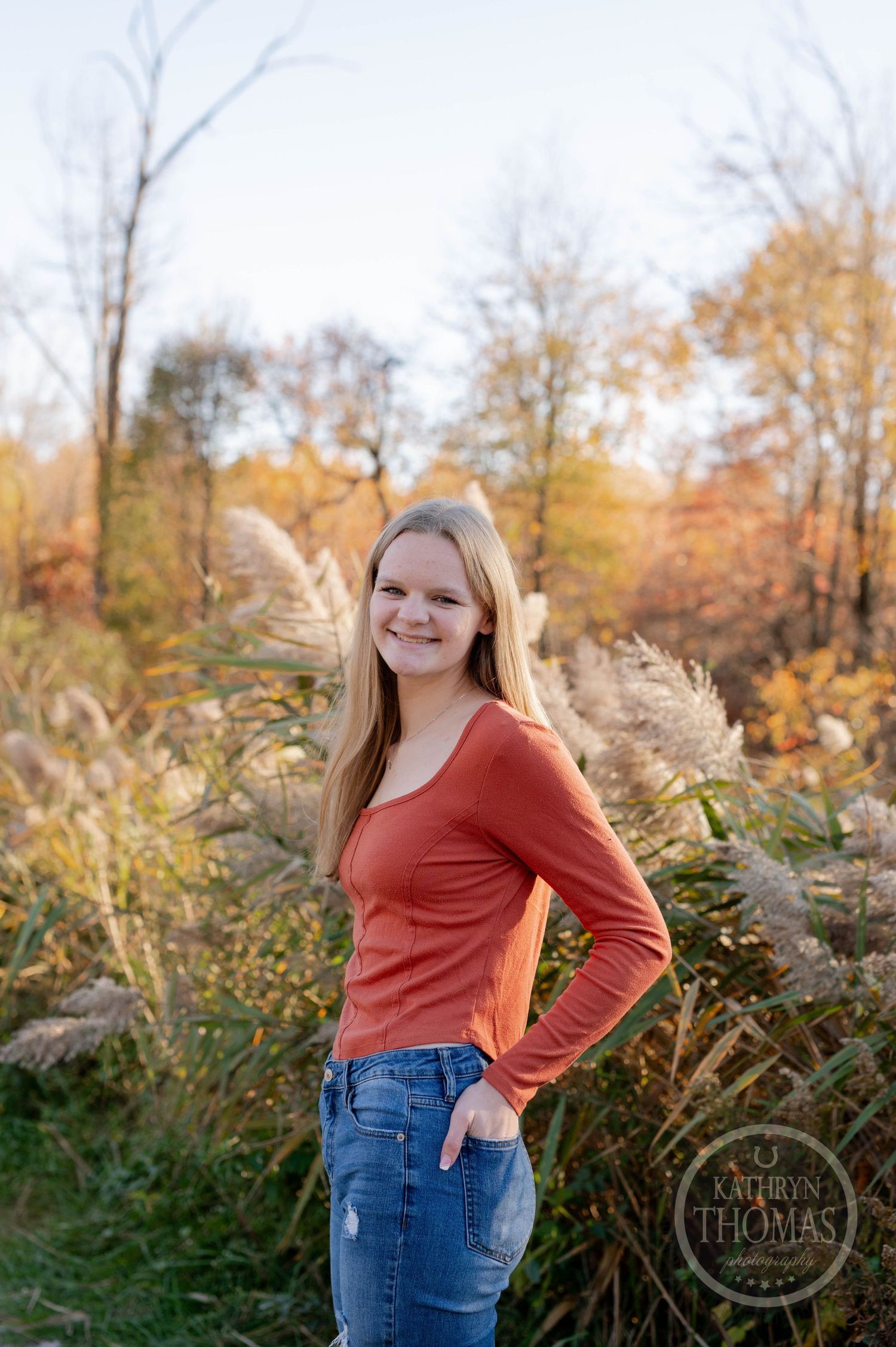 Person wearing orange sweater poses in front of vibrant fall foliage.