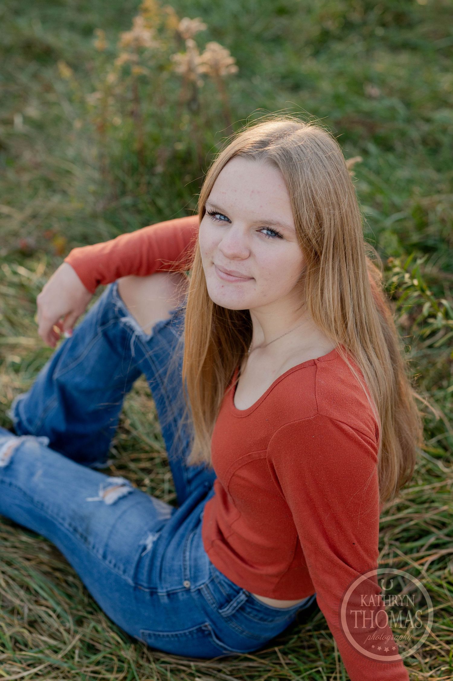 Casual outdoor portrait of someone in an orange sweater and jeans sitting in tall grass during golden hour.