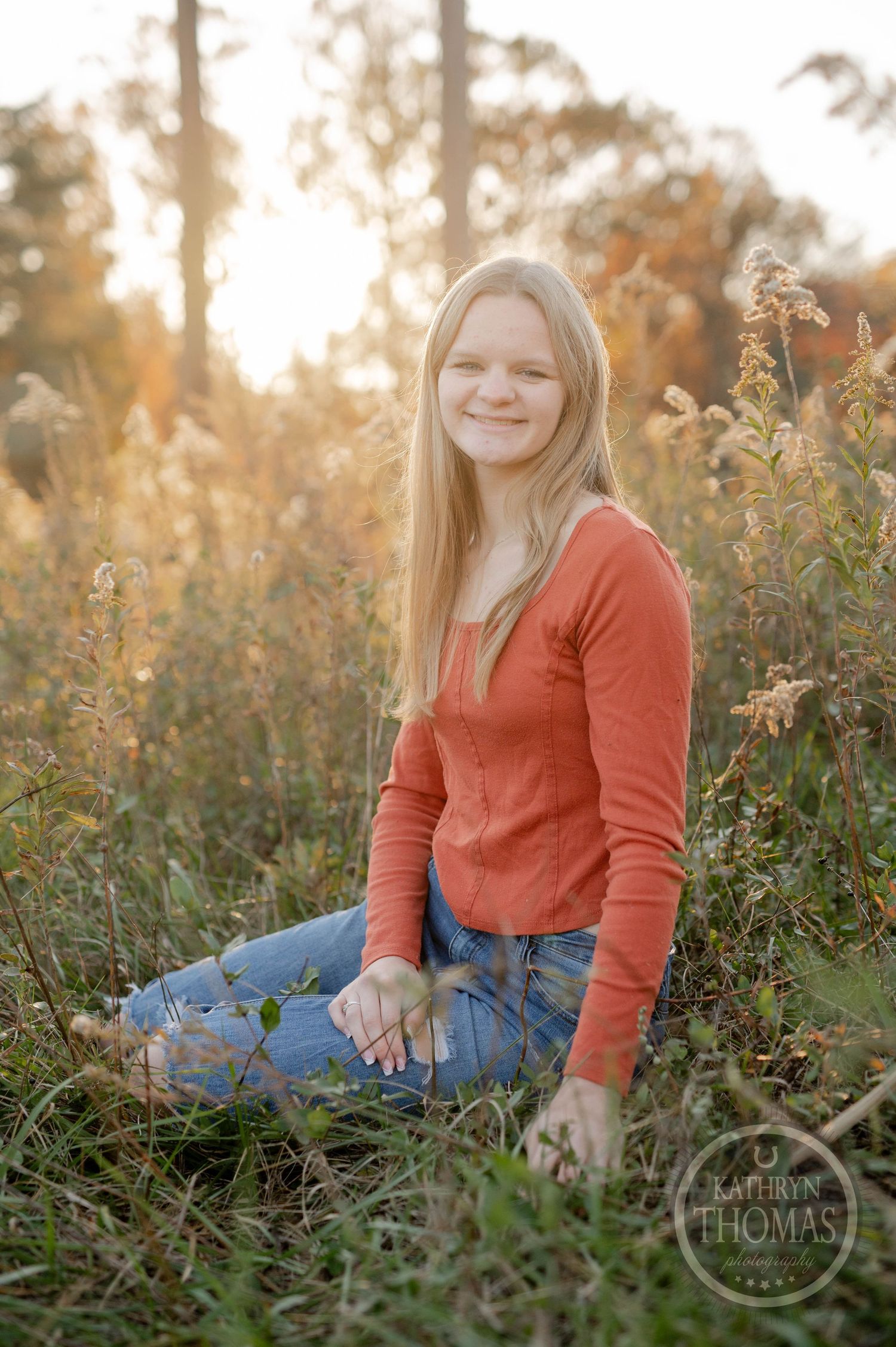 Person sits in tall grass wearing orange sweater during golden hour with fall trees behind.
