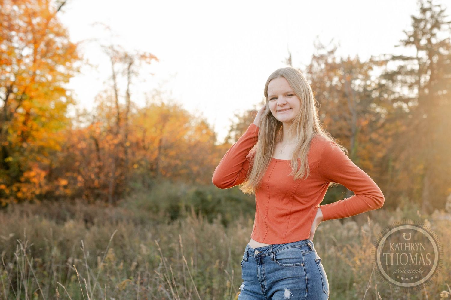 Person poses casually in orange top against golden fall foliage background.