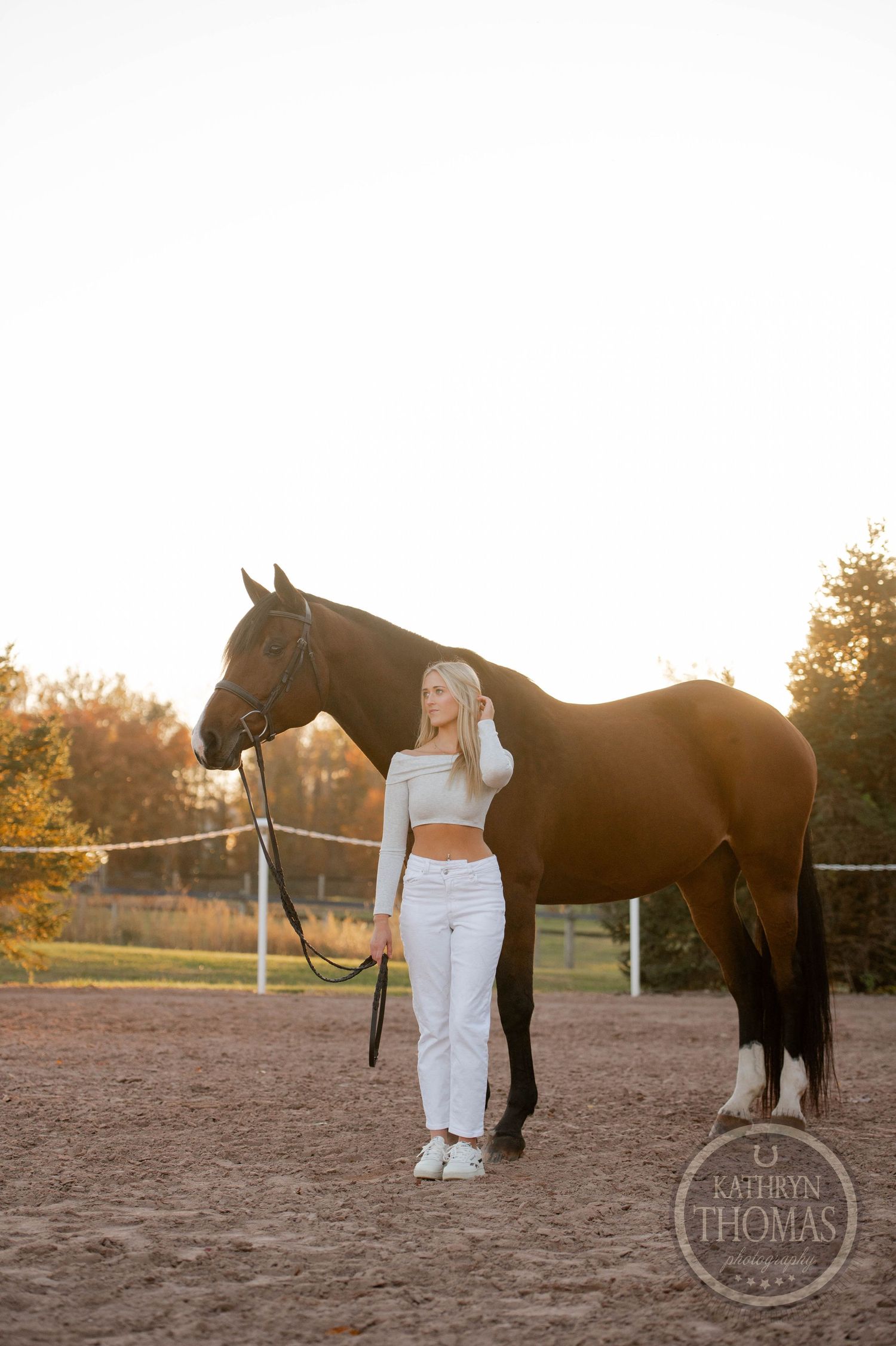 A person in white attire stands with a dark horse in a paddock at sunset.