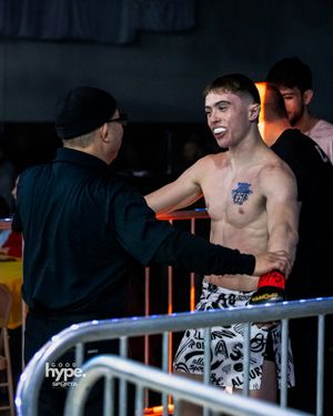 A shirtless fighter with a tattoo on his chest interacts with someone in black at the edge of a fighting cage.