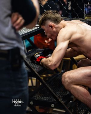 MMA fighter rests against cage fence during intense match with shirtless torso visible.