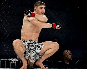 MMA fighter in black and white patterned shorts crouches in fighting stance inside octagon cage during competition.