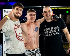 Three people pose together after a Muay Thai fight with one shirtless fighter in the middle celebrating in the ring.