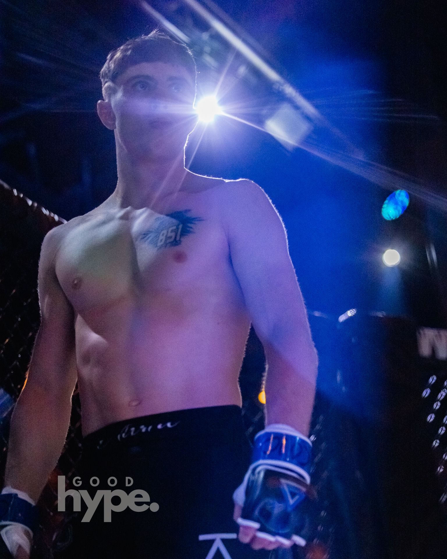 A shirtless MMA fighter in boxing gloves performing during a dark, dramatically lit fight event with blue lighting.