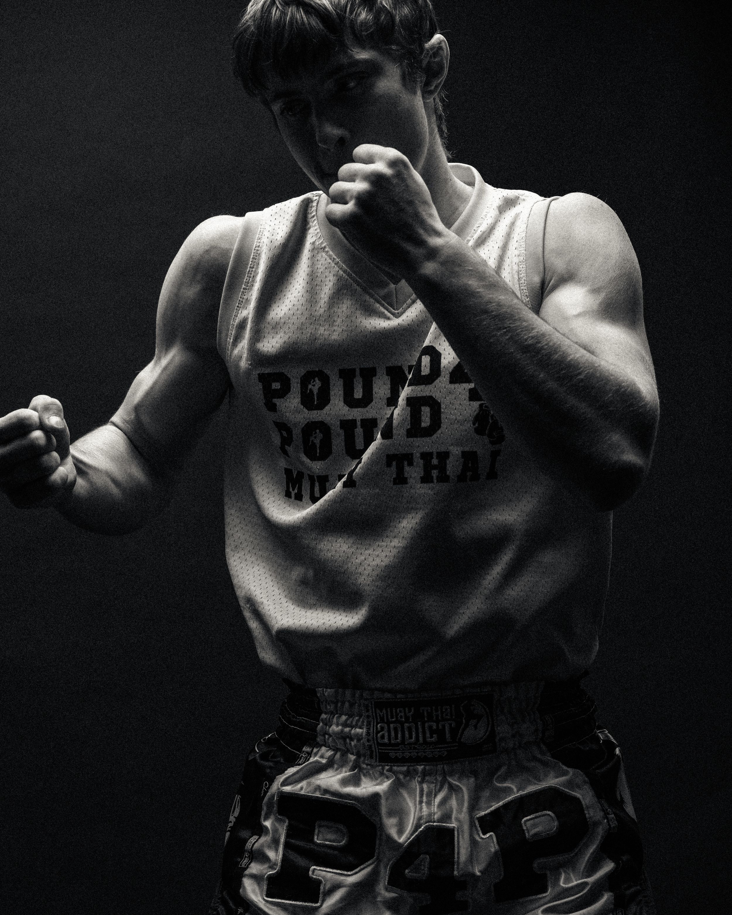 Athlete in a sleeveless tank top and boxing shorts assumes a fighting stance in dramatic black and white lighting.