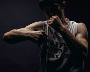 Dramatic low key portrait of muscular person in tank top wiping face against dark background.