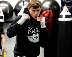 A boxer in a black Rush Down hoodie practices fighting stance with wrapped hands in front of punching bags at a gym.