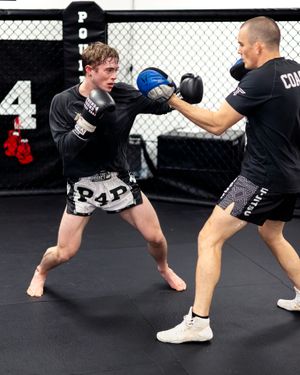 Two fighters sparring with protective gear during a Muay Thai training session in an MMA gym with chain-link fencing background.