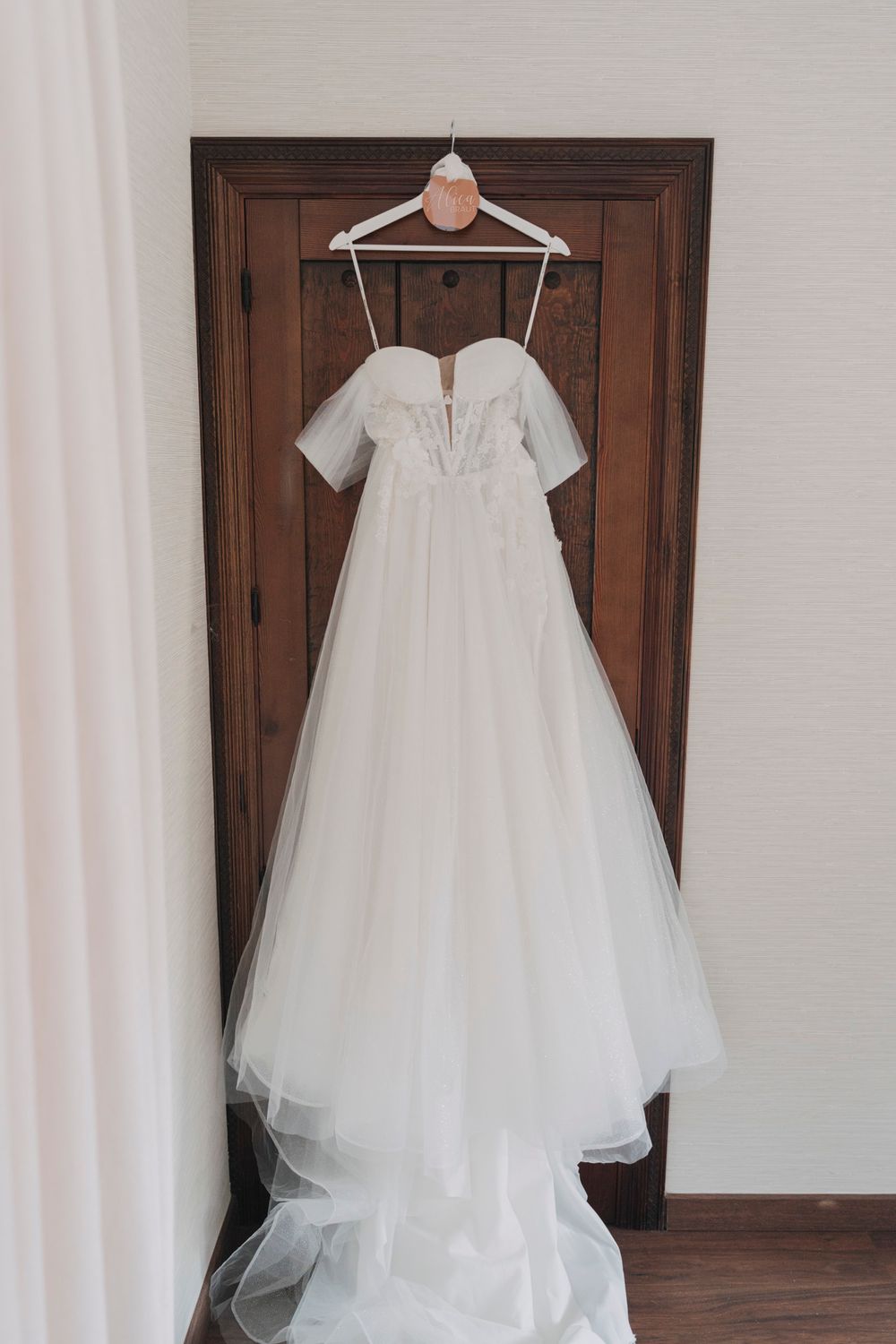 A white wedding dress with short sleeves and tulle skirt hangs on a wooden door against a light curtain backdrop.