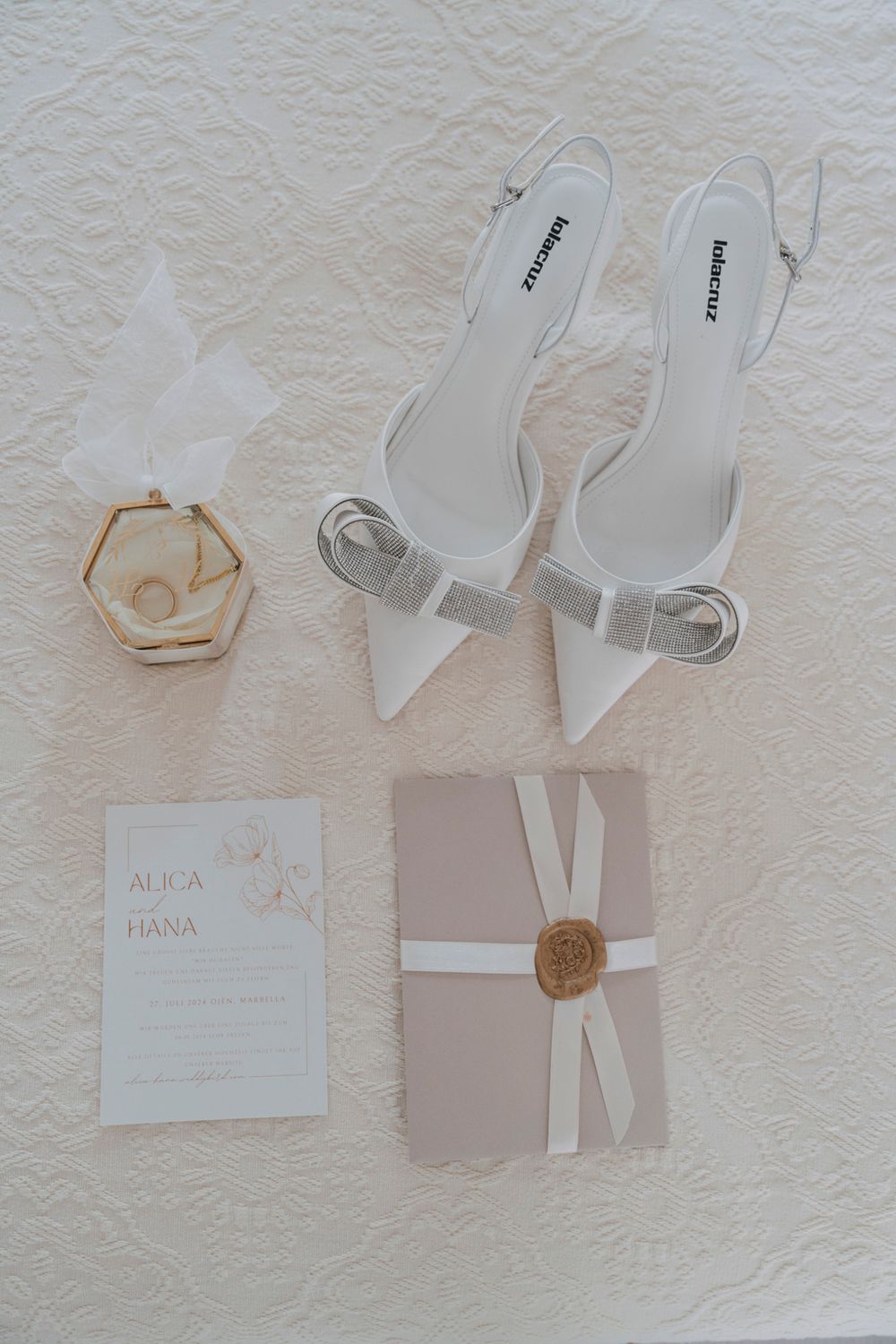 Elegant white bridal accessories including pointed slingback heels, hexagonal jewelry box, and invitation suite on cream background.