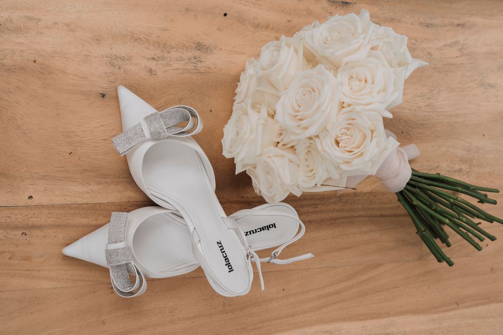 A pair of elegant white bridal shoes next to a delicate bouquet of cream-colored roses on a wooden surface.