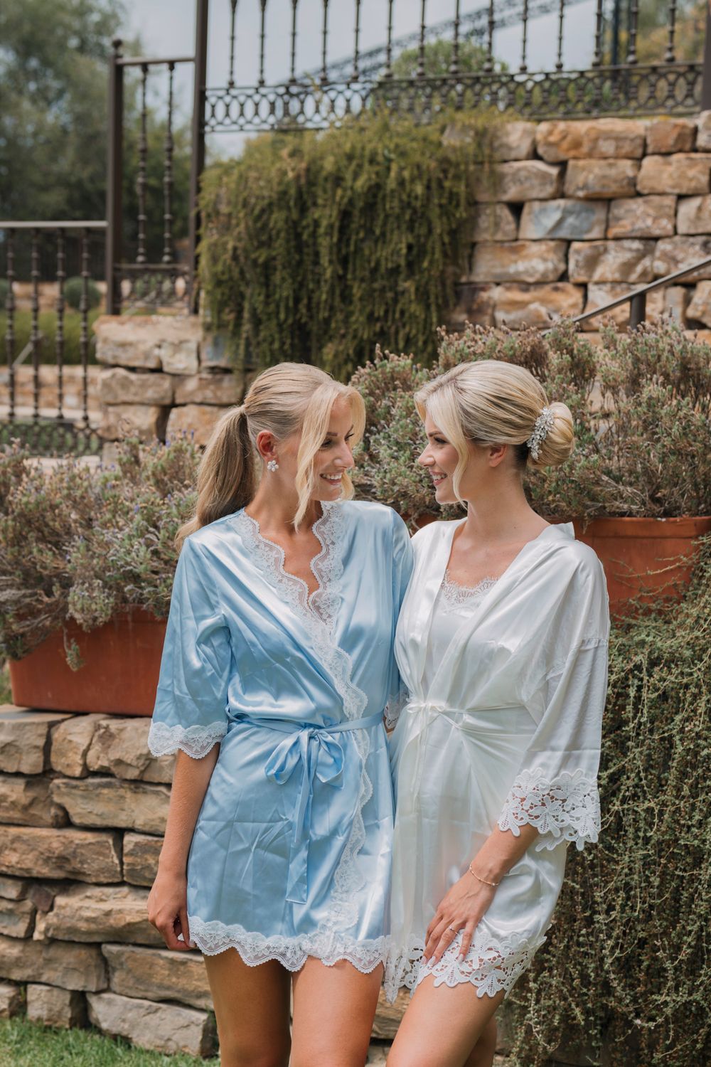 Two matching satin robes against stone wall and garden backdrop at wedding venue.