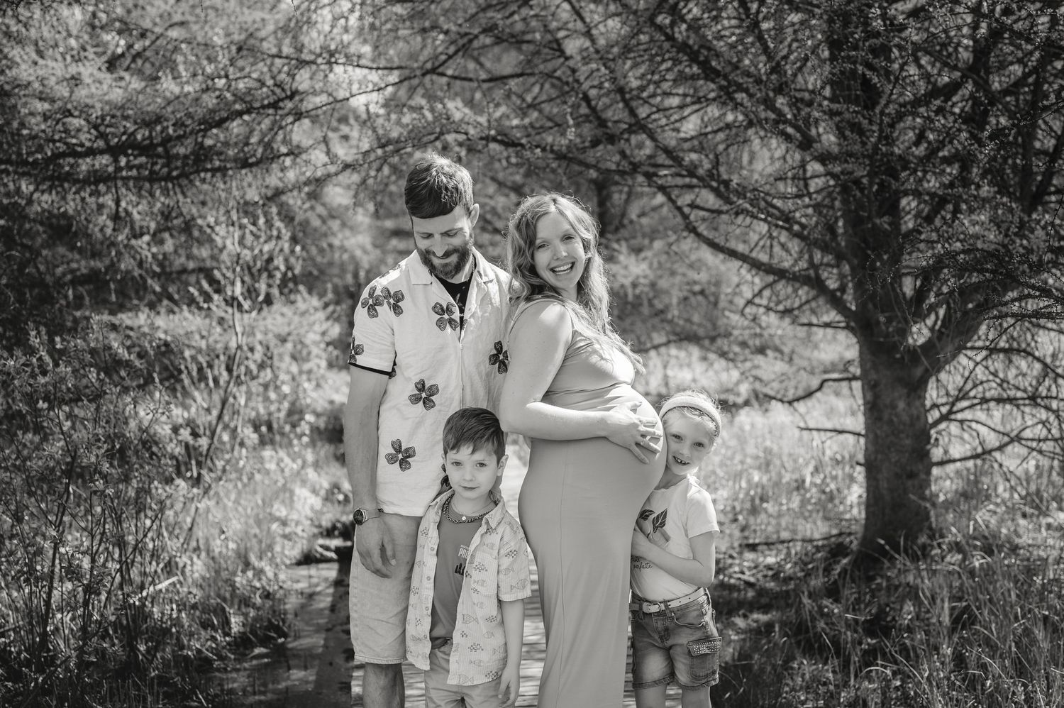 Black and white family portrait in wooded setting during maternity photoshoot.