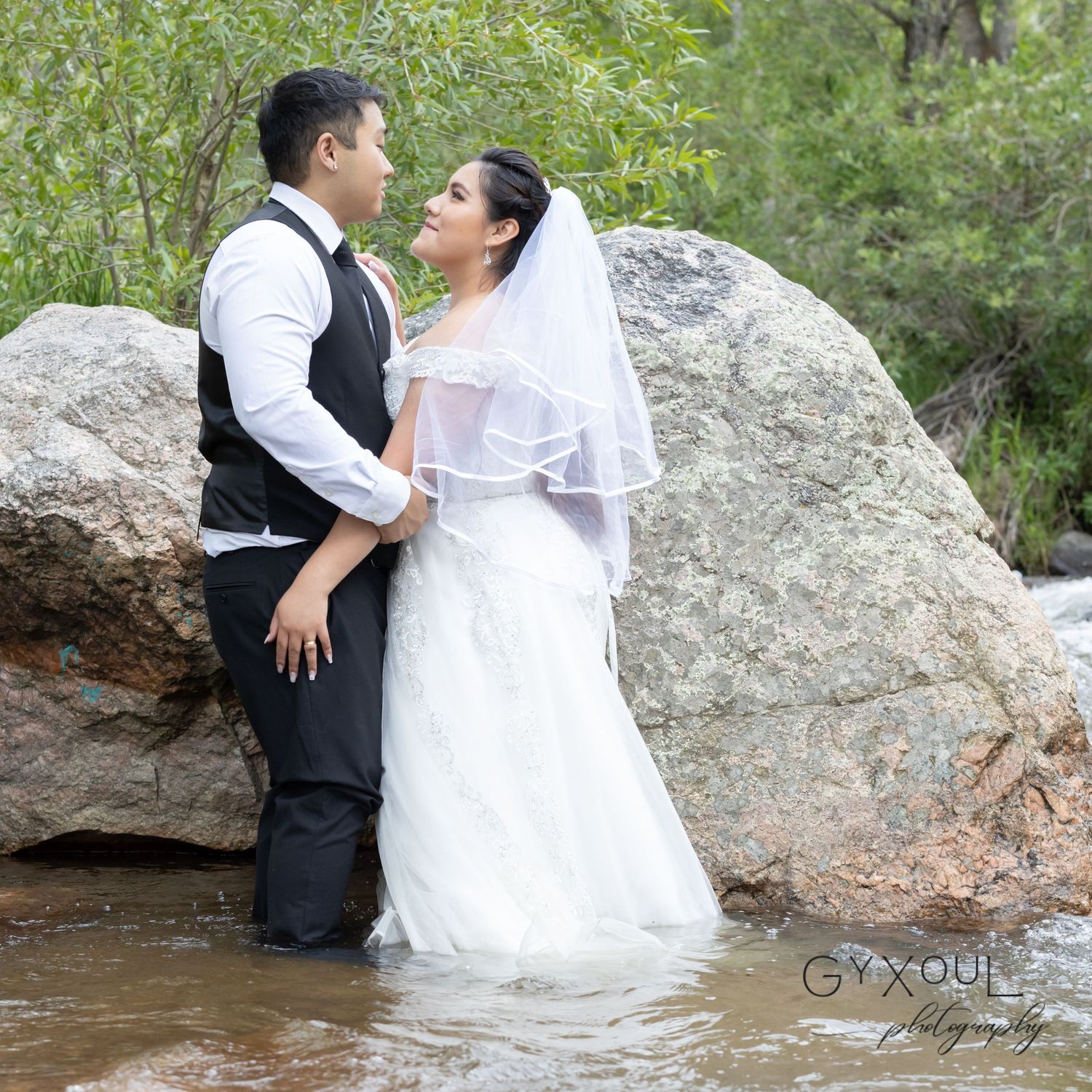 A newlywed couple poses in creek water against large rocks in a romantic outdoor setting.