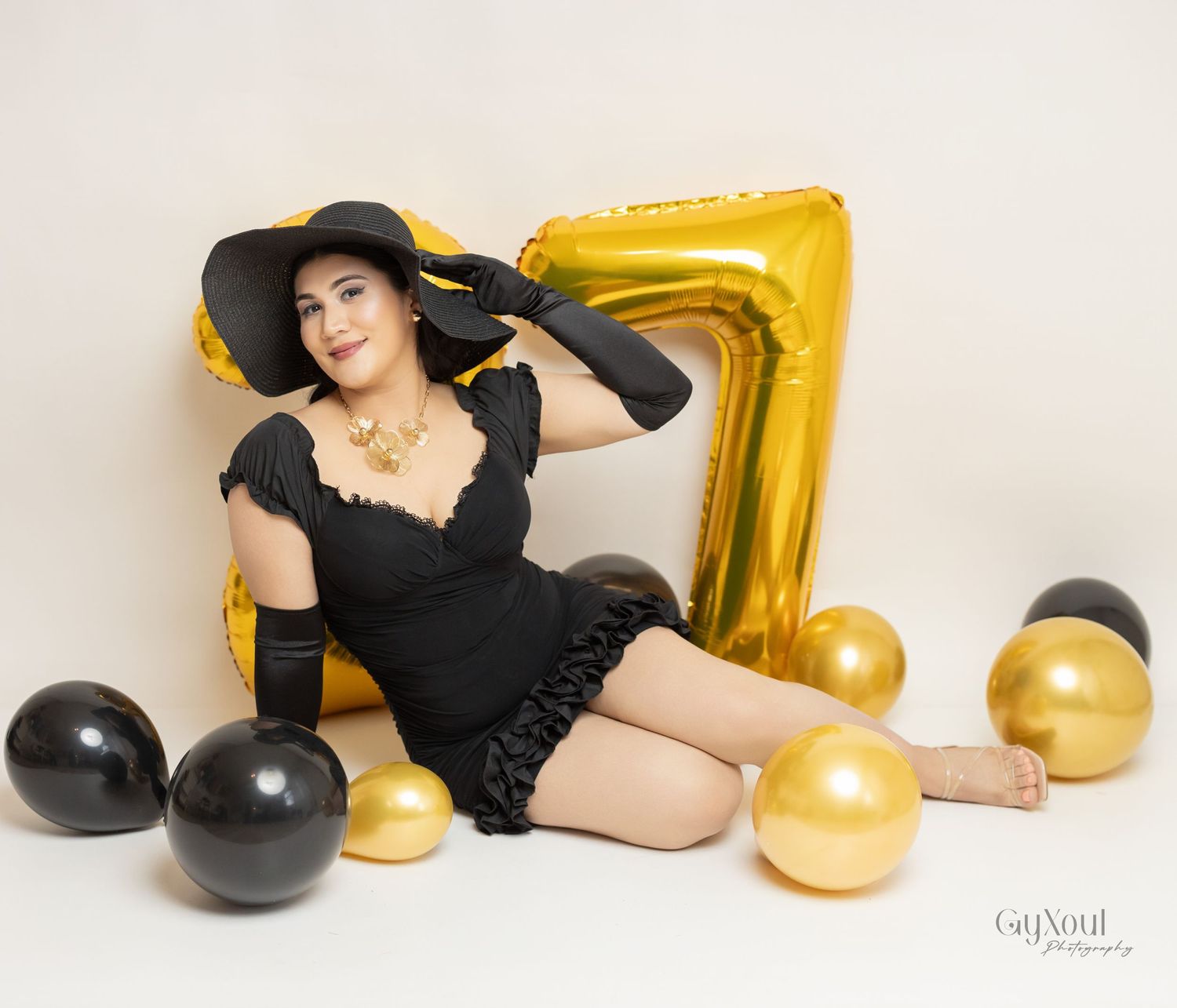 Retro glamour Vintage-inspired birthday photoshoot in black dress and hat against gold '7' balloon and party decorations.