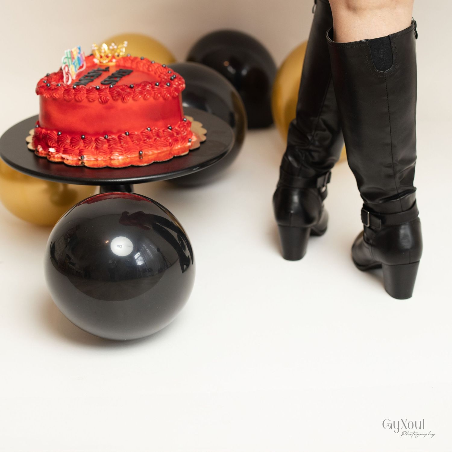 Red birthday cake on black plate with gold and black balloon decorations and black boots visible.