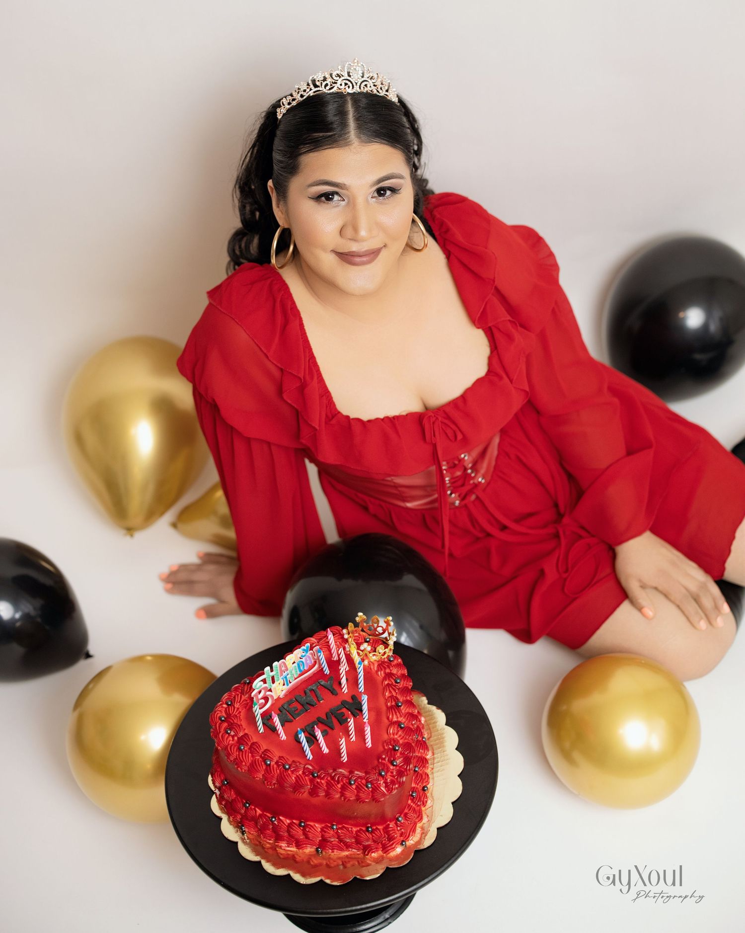 Birthday celebration setup for photo shoot with red cake, gold and black balloons, and sparkly tiara.