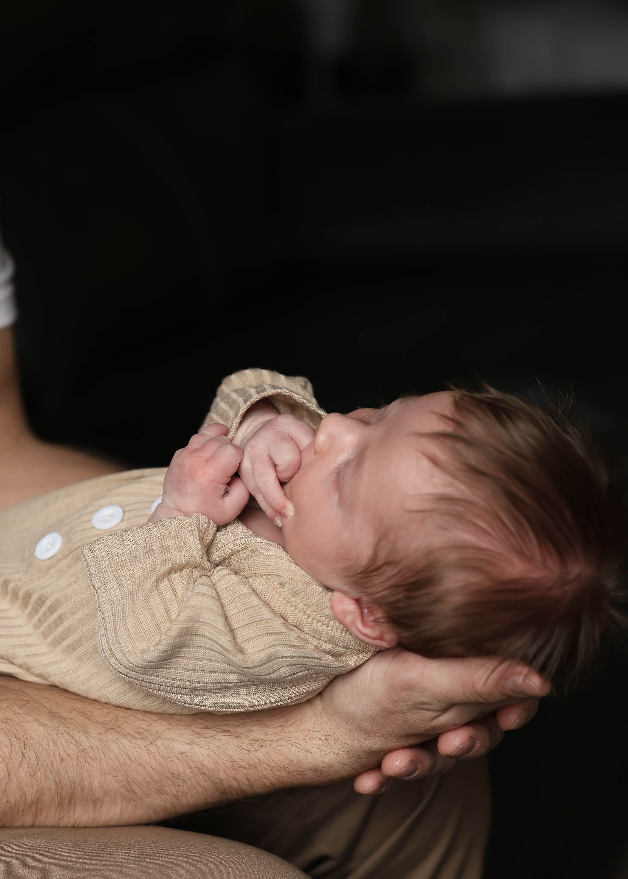 A baby in a beige knitted sweater lying in supportive hands against a dark background.