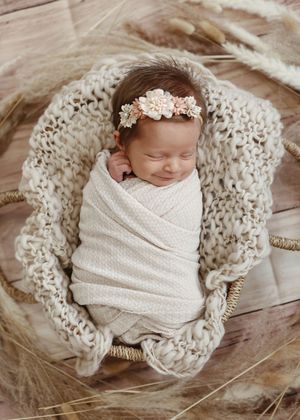 Sleeping newborn wrapped in white swaddle on knit blanket with floral headband.