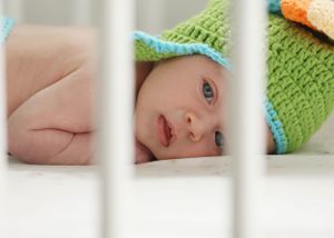 A baby wearing a mint green knit hat peeks through white crib bars.