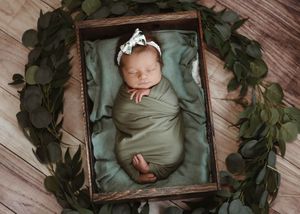 Newborn photography series with baby wrapped in olive green fabric surrounded by greenery and wooden frames.