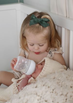 A toddler with a green bow feeding a baby doll in a white nursery setting.