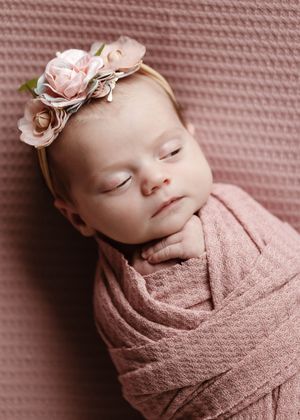 Peaceful newborn wrapped in pink fabric with floral crown against textured rose background.