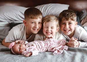 Three smiling children lying on a bed with a newborn baby wrapped in a pink floral swaddle blanket.