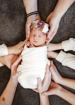 Newborn baby wrapped in white swaddle with floral headband, cradled by multiple family hands on gray background.