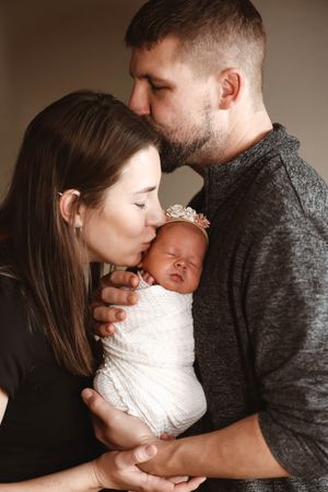 New parents kissing their swaddled newborn baby girl wearing a floral headband.