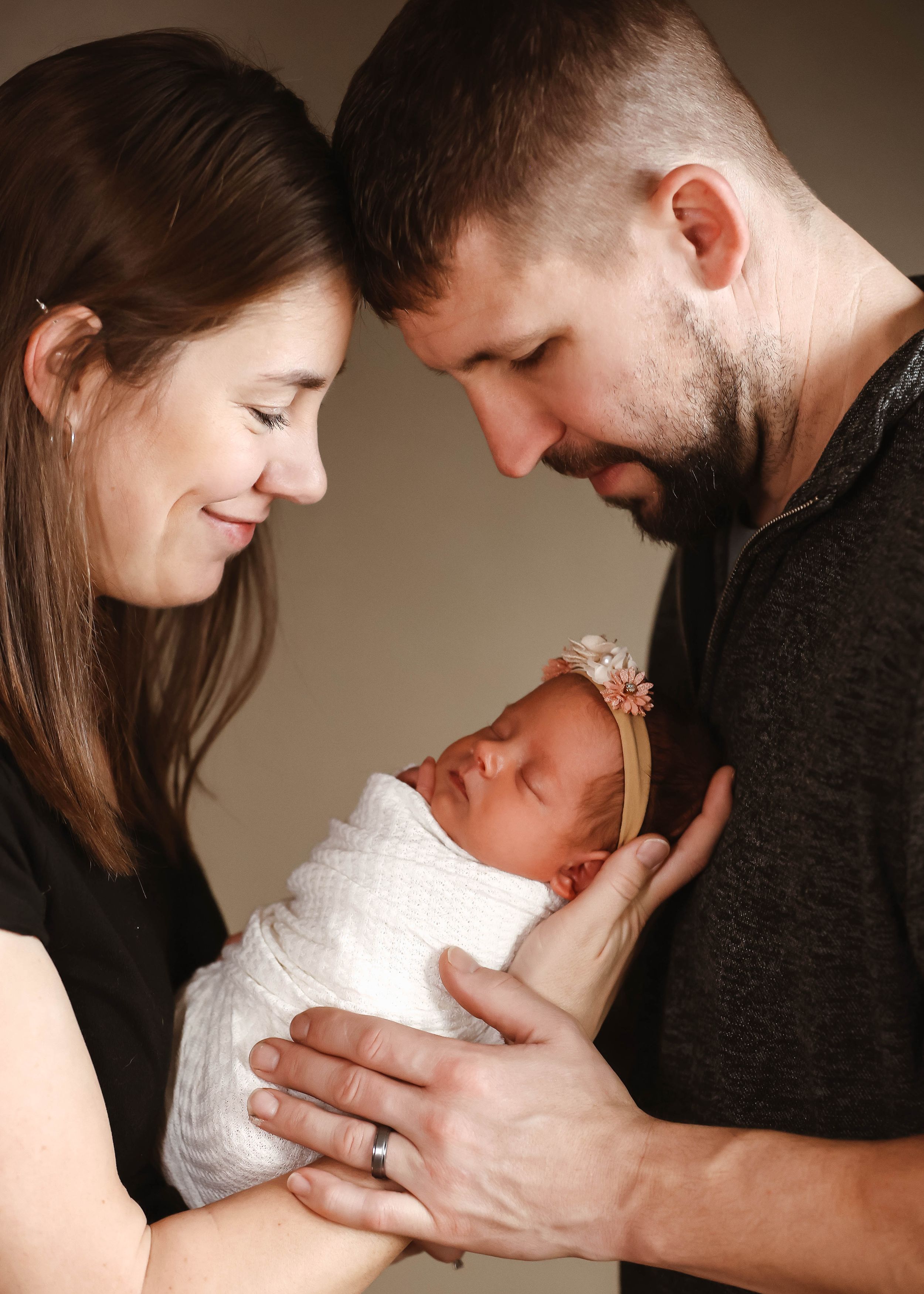 Parents lovingly gaze at their swaddled newborn baby girl wearing a floral headband in a tender family portrait.