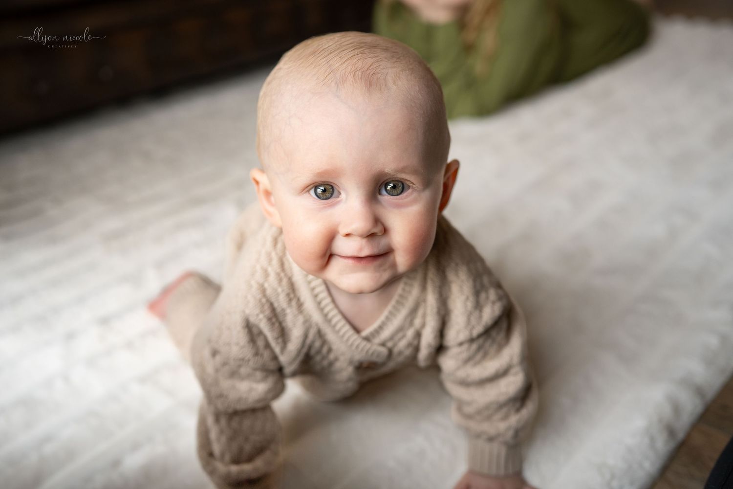 Happy baby wearing a beige sweater lying on white bedding while smiling at camera.