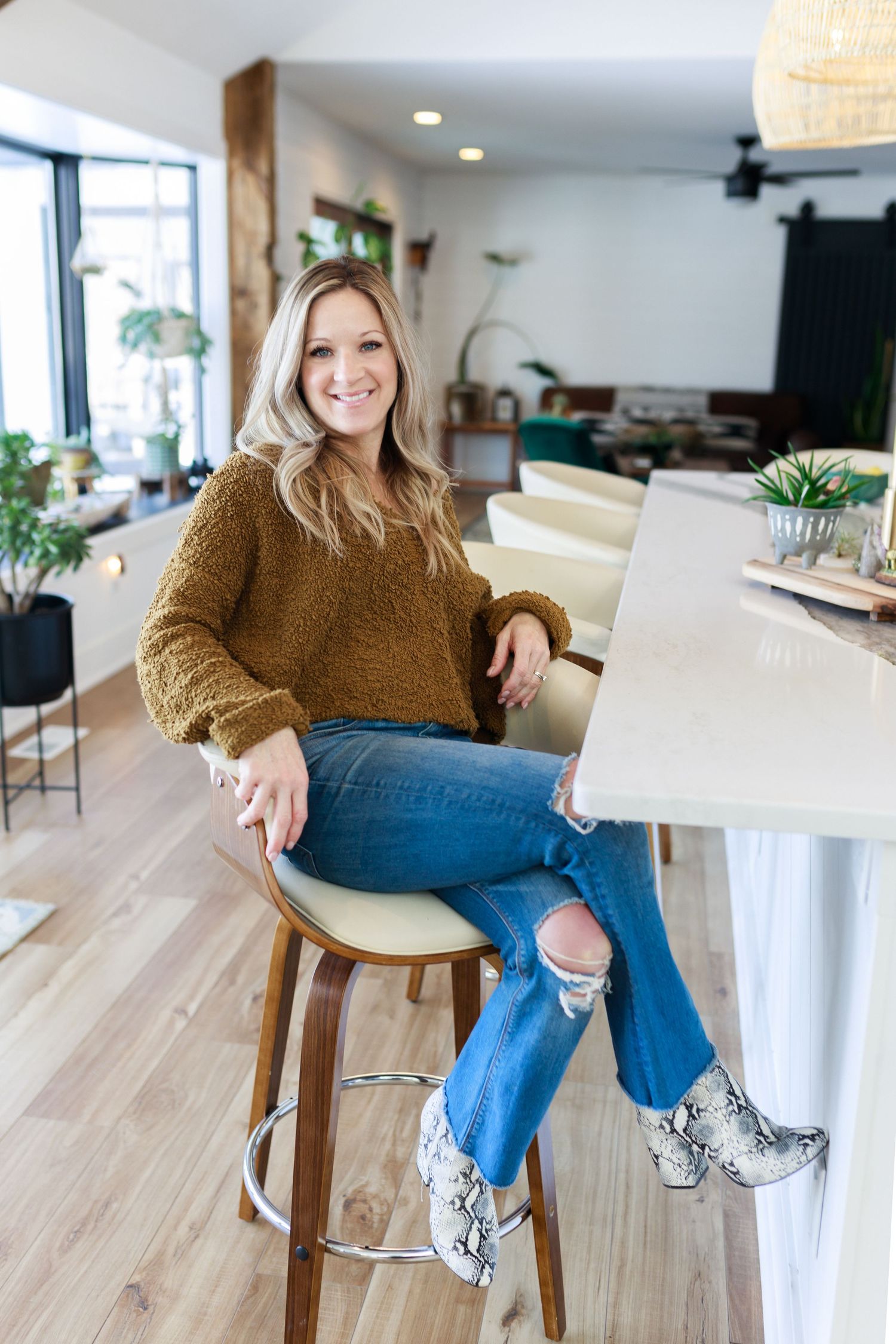 Valparaiso business owner in brown knit sweater and ripped jeans sits casually on bar stool in modern home interior with hardwood floors.