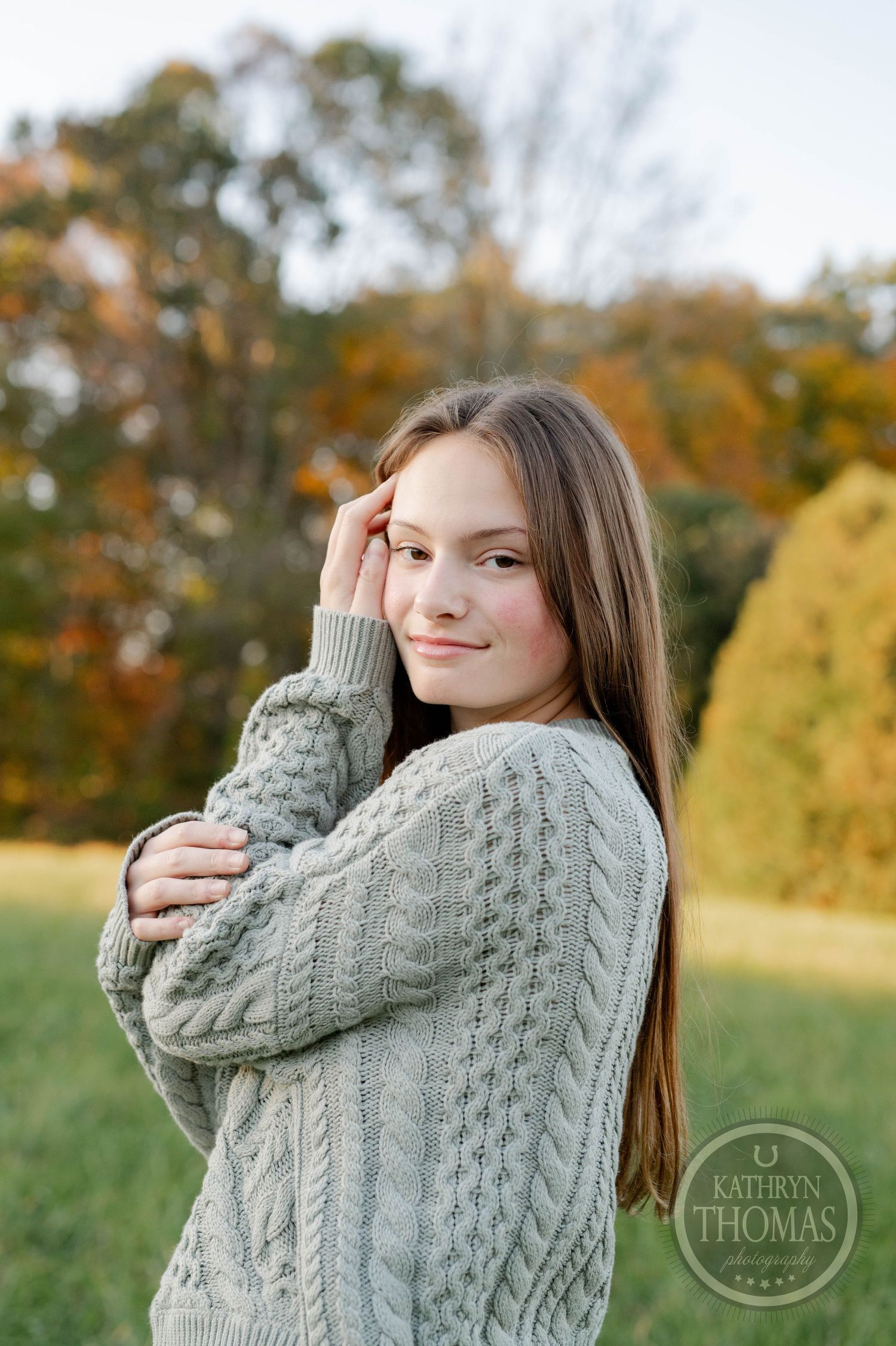 A cozy autumn portrait features someone in a chunky gray cable knit sweater against falling foliage.