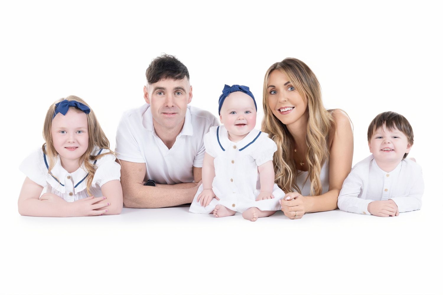 A family of five wearing white outfits lies together on the floor against a white background.