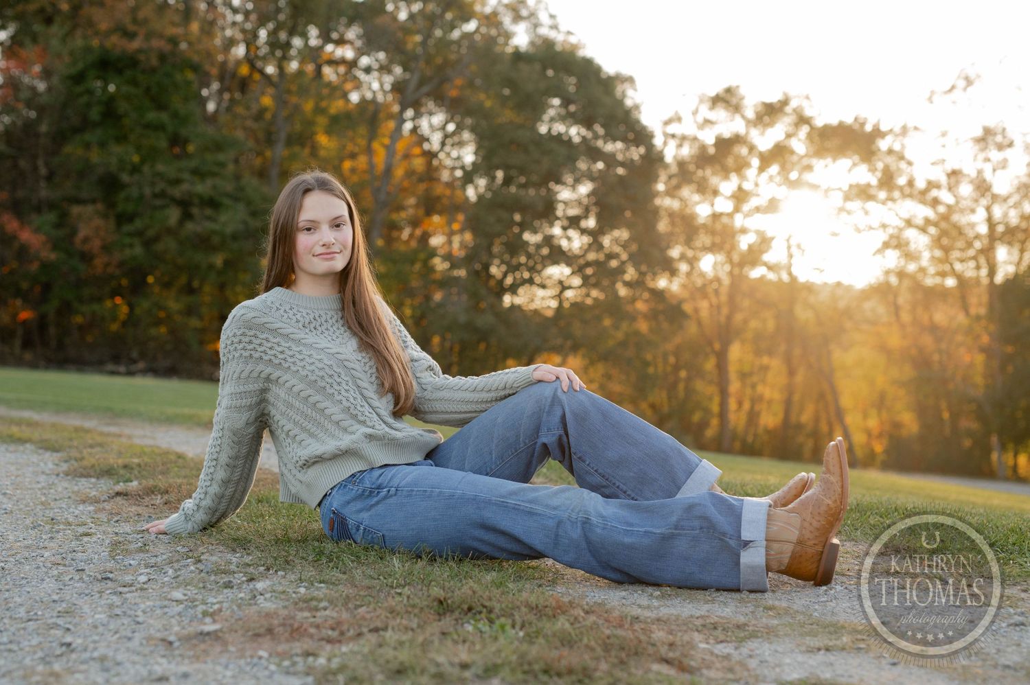 Person in gray sweater relaxing on dirt path during golden hour sunset with autumn trees in background.