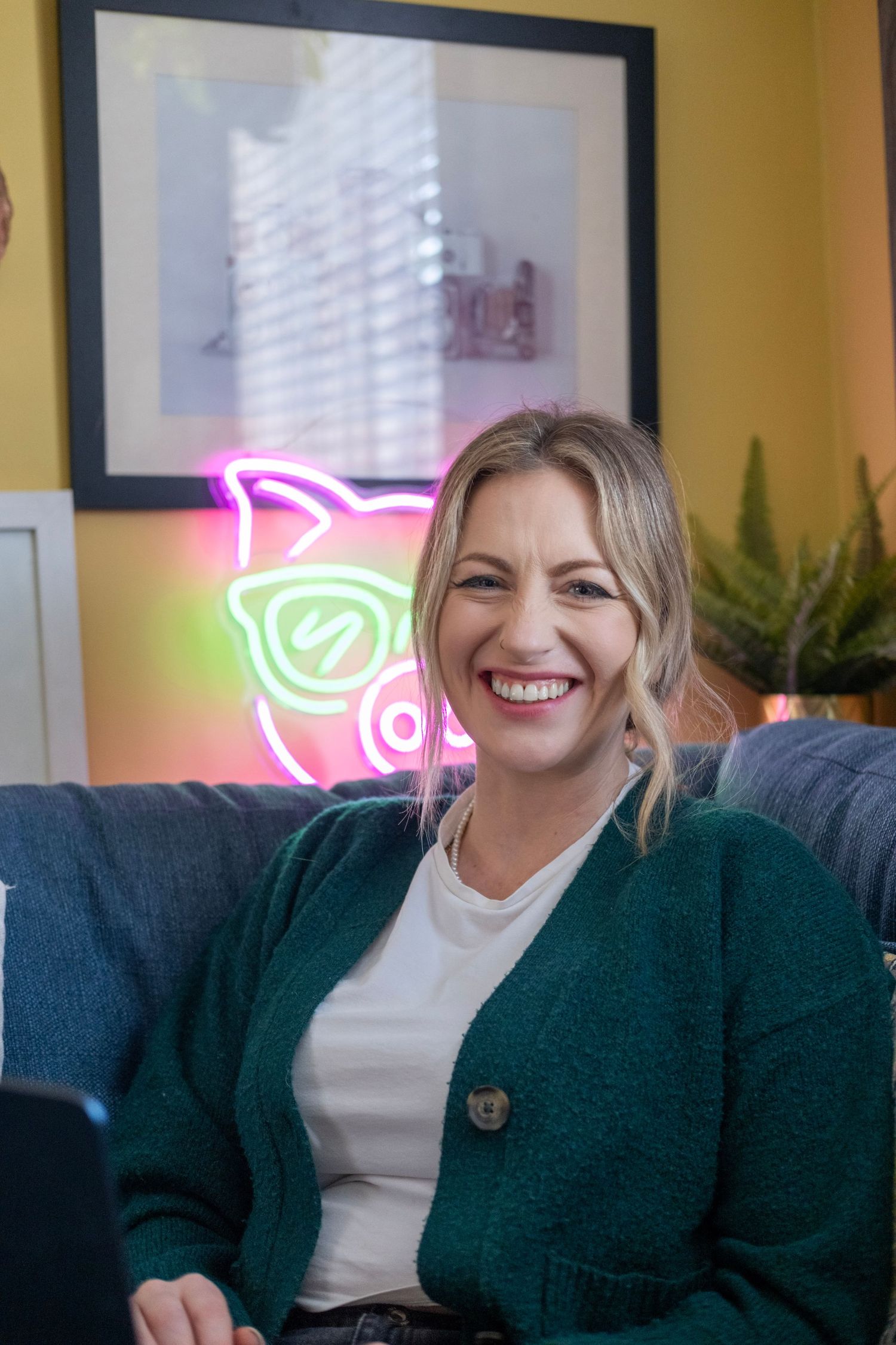 Person smiles warmly on blue couch with neon rose sign glowing in background.