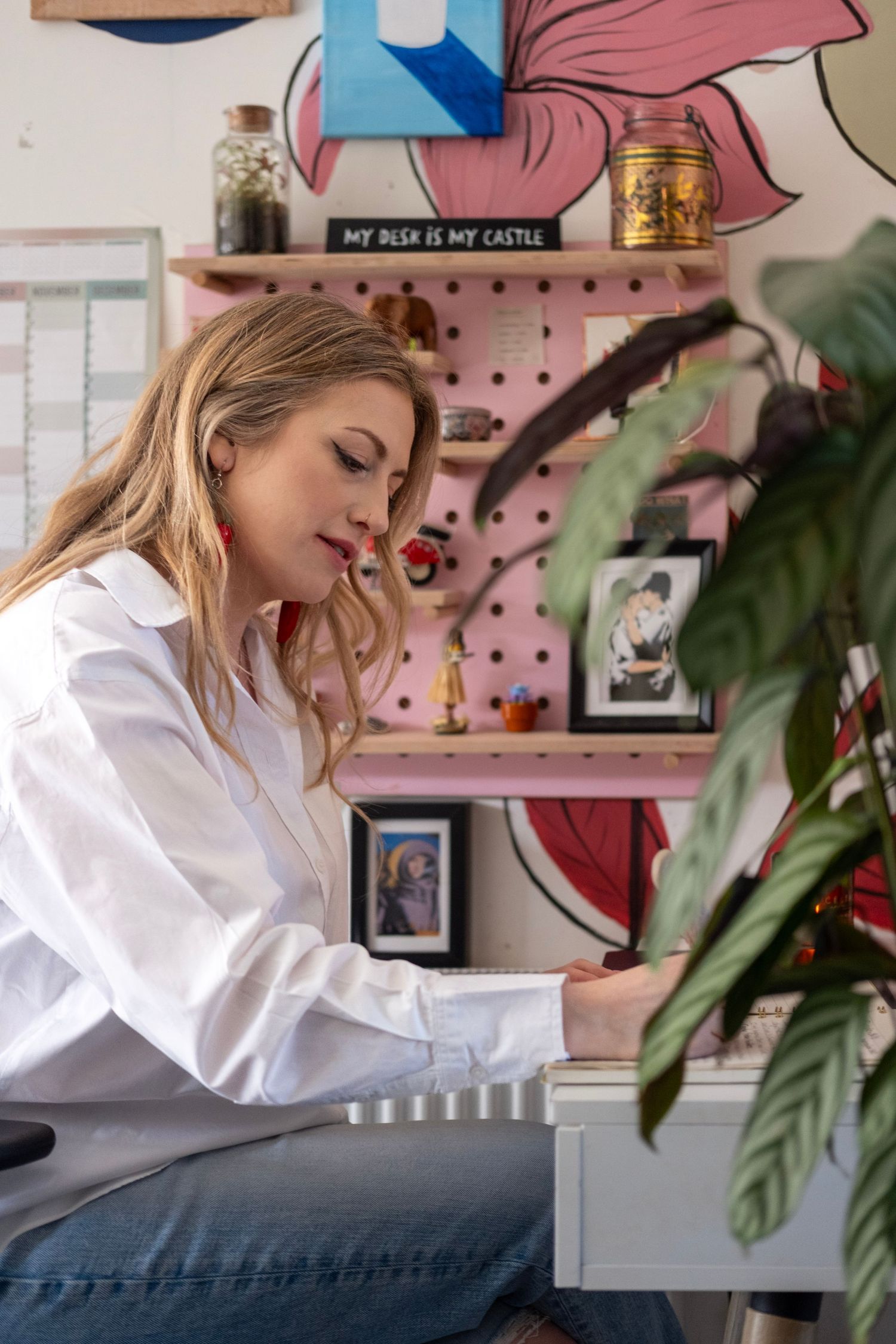 Person writing at desk with pink pegboard wall and indoor plants in creative workspace.