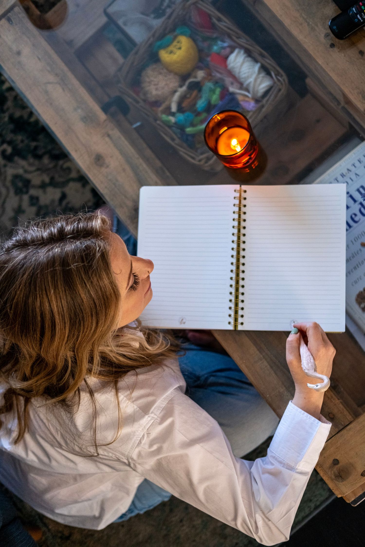 Overhead view of person writing in blank notebook with candle light on wooden surface.