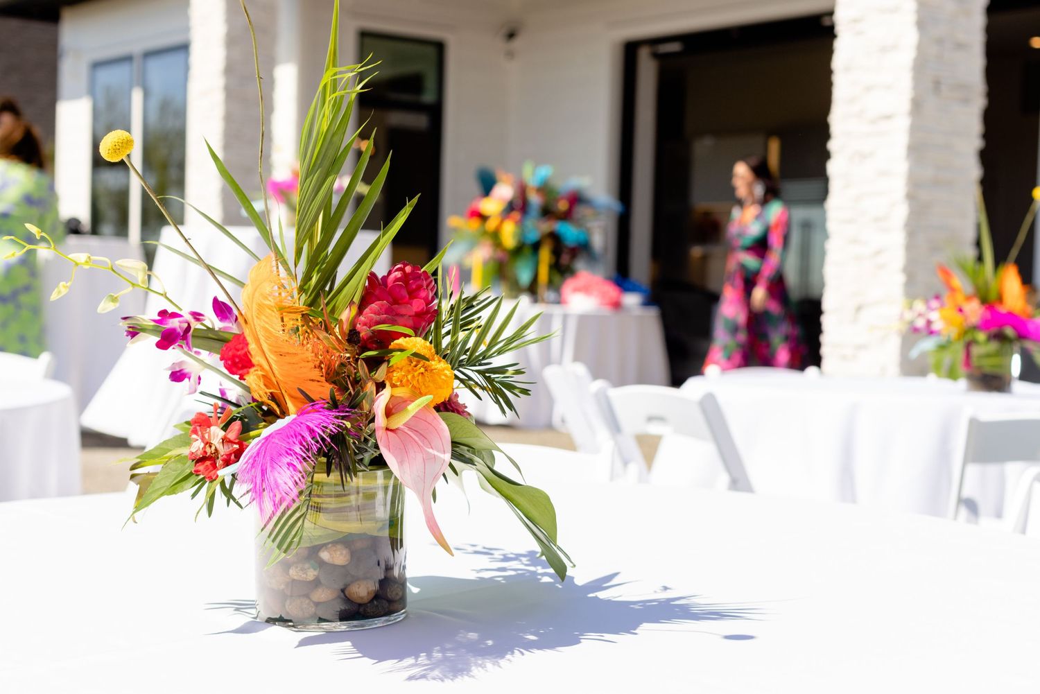 Colorful tropical floral centerpiece with bright pink and orange flowers on white tablecloth at outdoor event venue.