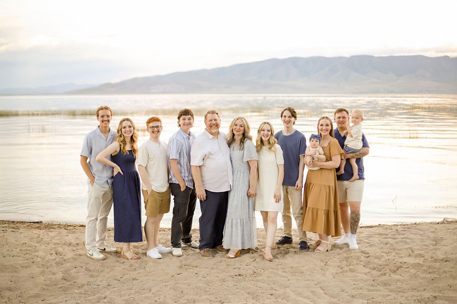 Group dressed in coordinated light colors stands together on a sandy beach at sunset with mountains in background.