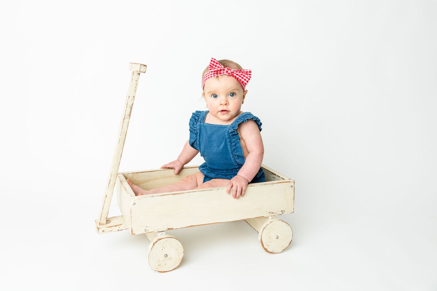 Baby in blue outfit and red headband sits in vintage white wooden wagon against white backdrop.