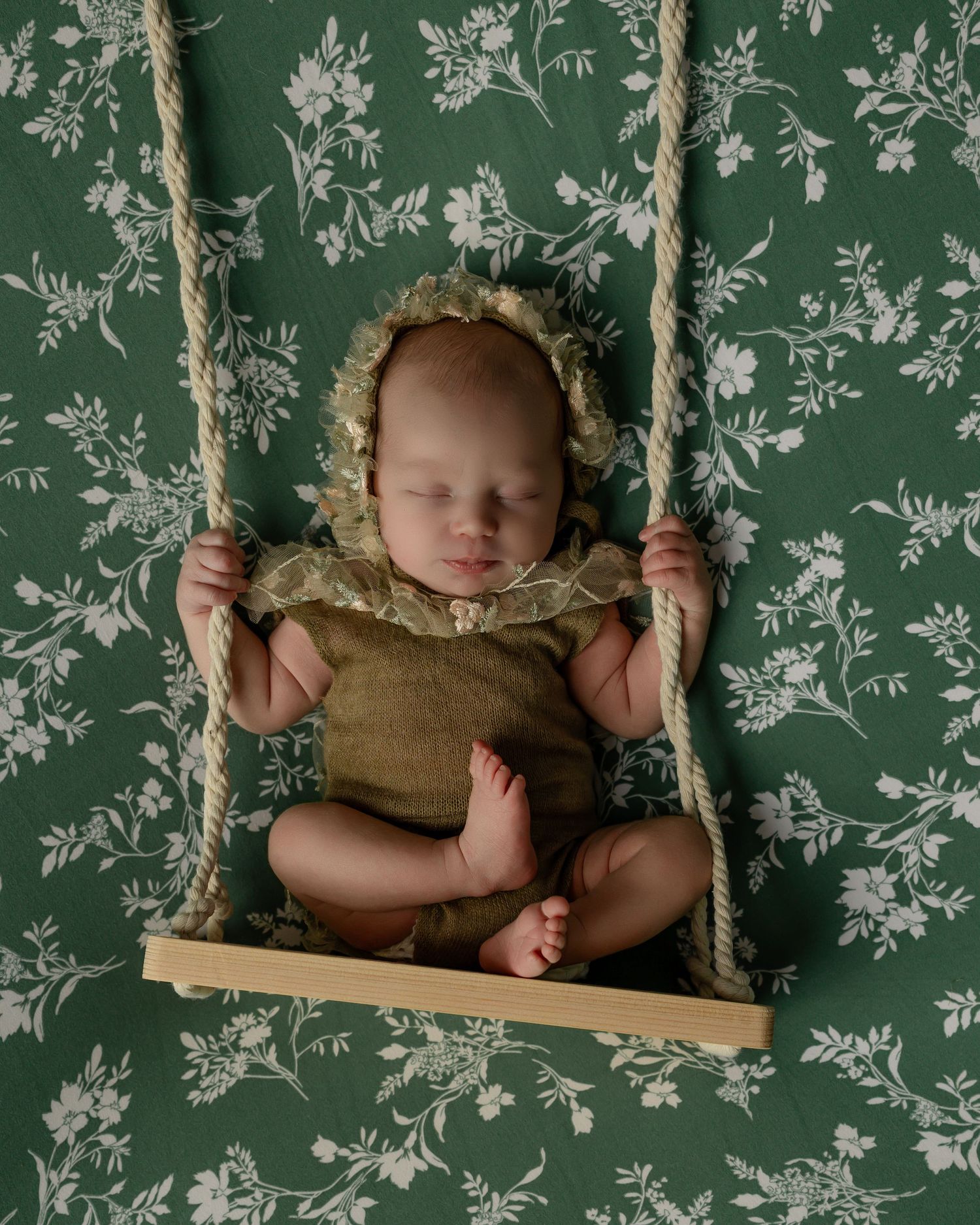 Sweet sleeping baby wearing olive bodysuit and bonnet on wooden swing against green and white floral wallpaper.