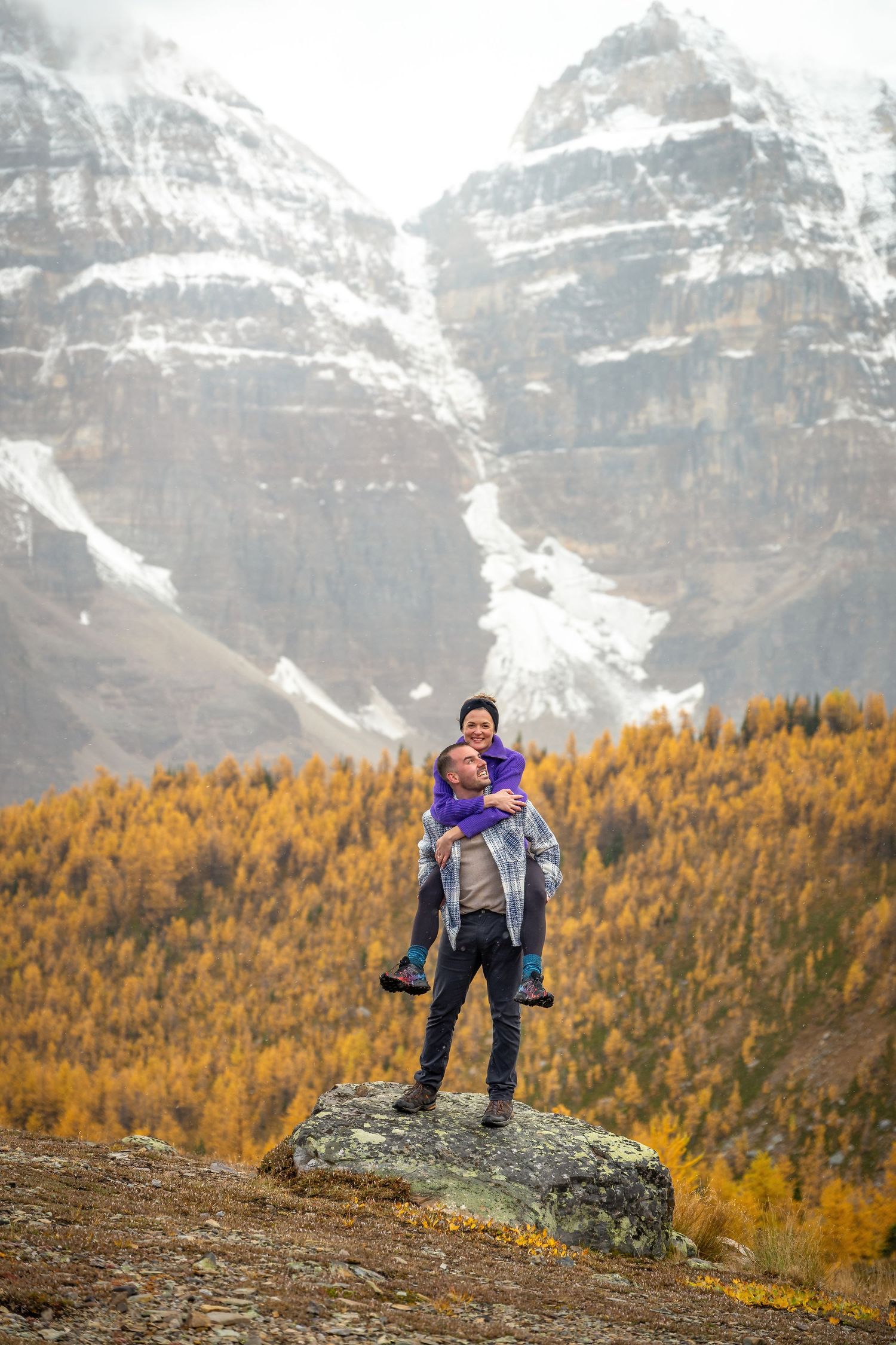 Two hikers stand triumphantly on mountain peak with autumn larches and snowy peaks behind.