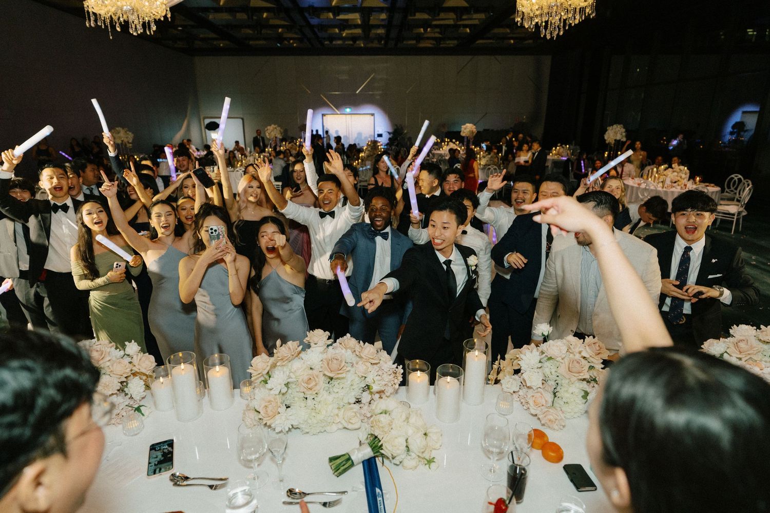Wedding guests raise their hands in celebration at an elegant reception with crystal chandeliers and decorated tables.